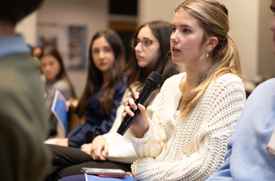 A young blonde woman in a white sweater sits in the crowd among peers and holds the microphone, poised to ask a question.