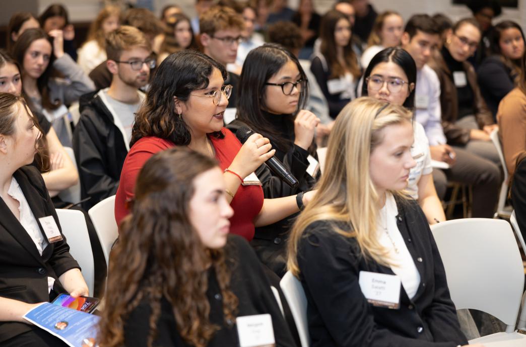 A young woman wearing glasses and a red top sits among a crowd of peers. She holds the microphone and is mid sentence.