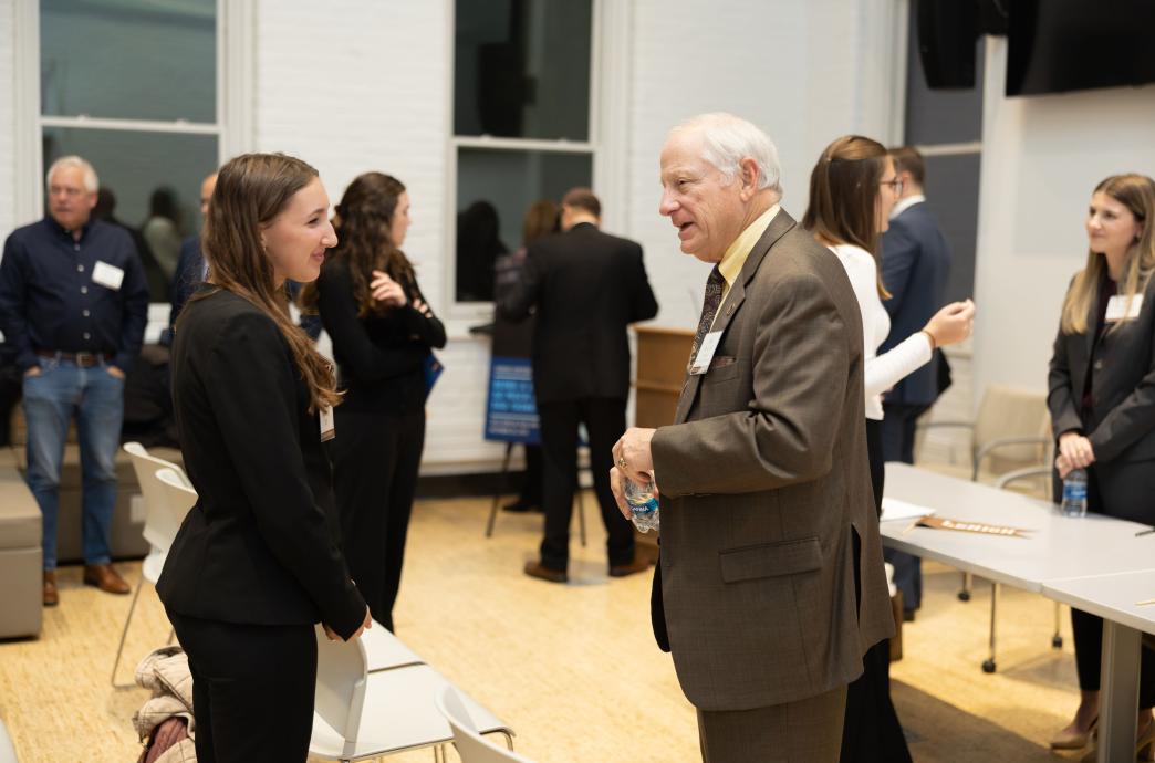 A young woman, dressed in all-black professional attire, chats to an older gentleman in a brown suit; people chat around them.