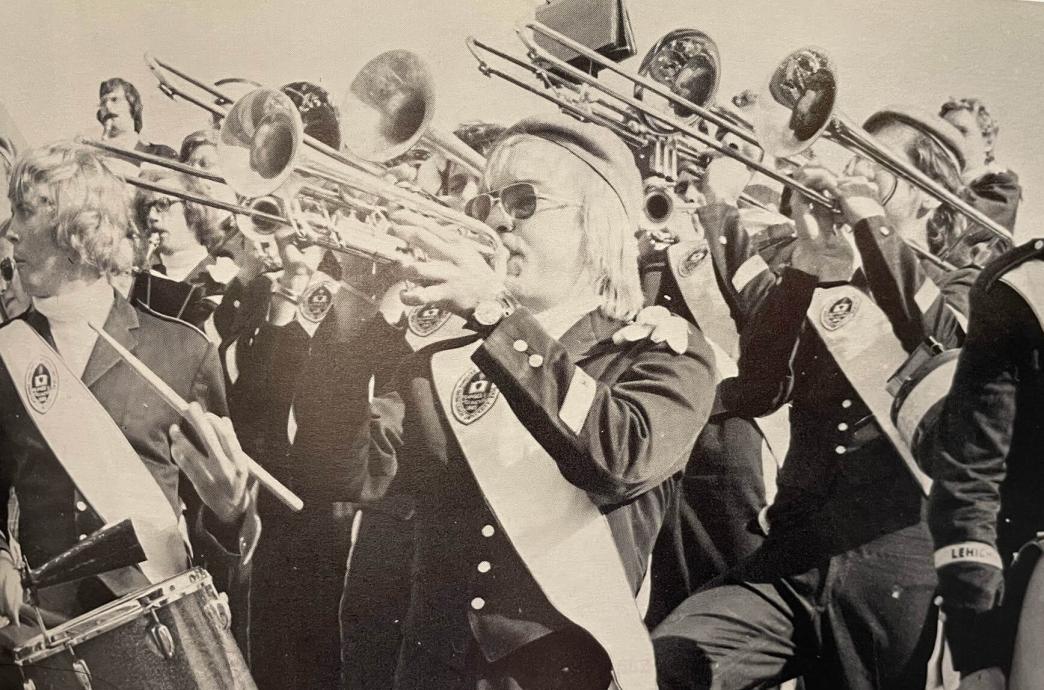 In a black and white photo, a marching band in full uniform is shown with focus on the horn section.