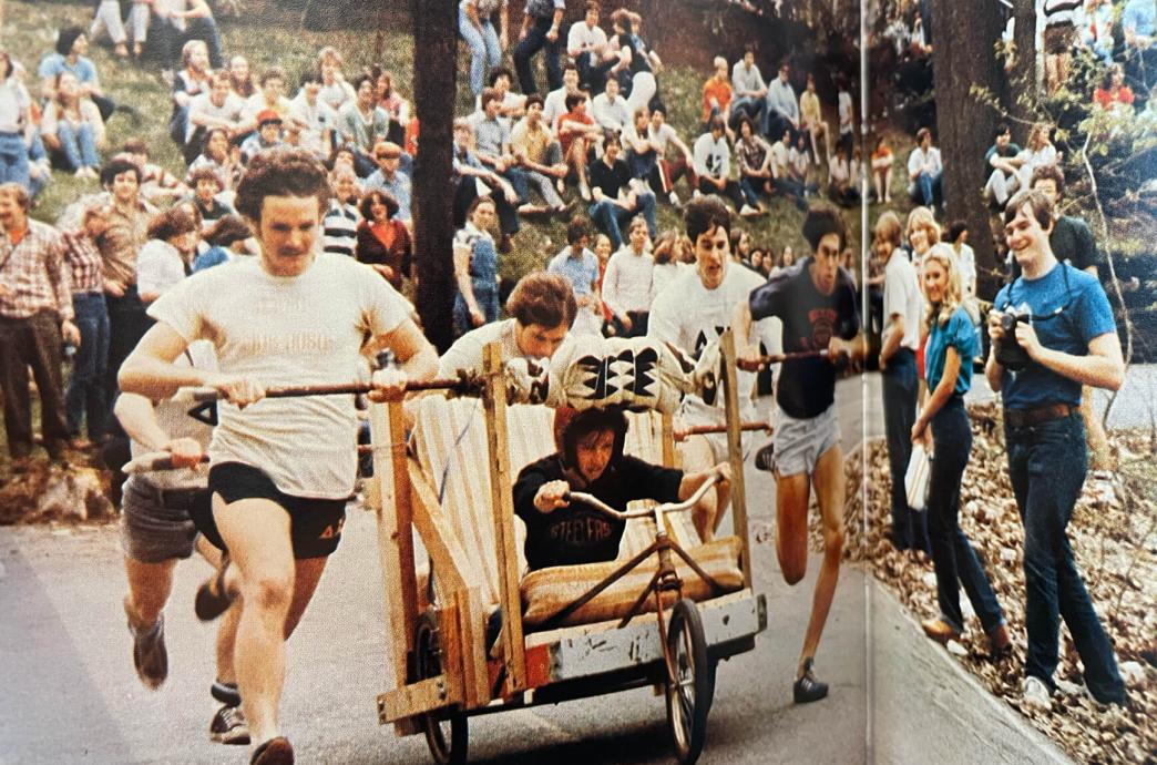 A vintage color photo shows a handmade car made of a bed frame and bicycle, pushed by students with one steering inside.