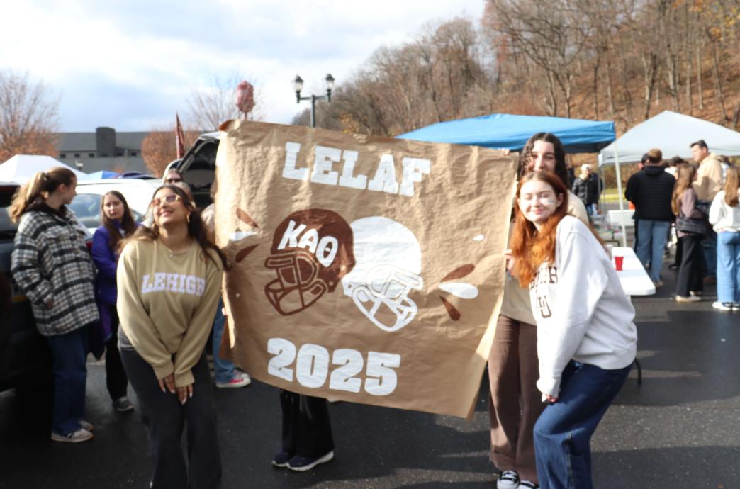 Three young women in Lehigh sweaters hold up a large, hand-painted sign that reads 'LeLaf 2025' with two brown and white helmets.