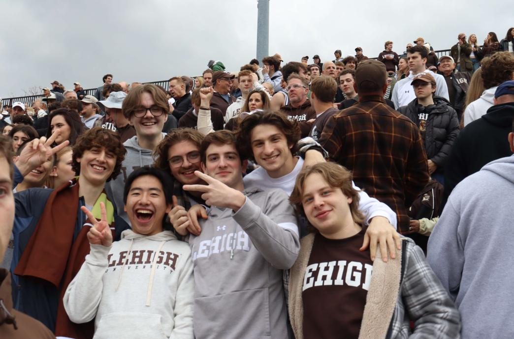 Amid a sea of fans, a group of seven young men smile at the camera while wearing Lehigh merch; three of them hold up peace signs.