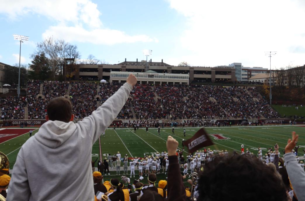 Fans cheer and fill the stadium bleachers. In the foreground, a young Lehigh fan cheers while a Lehigh flag is waved.