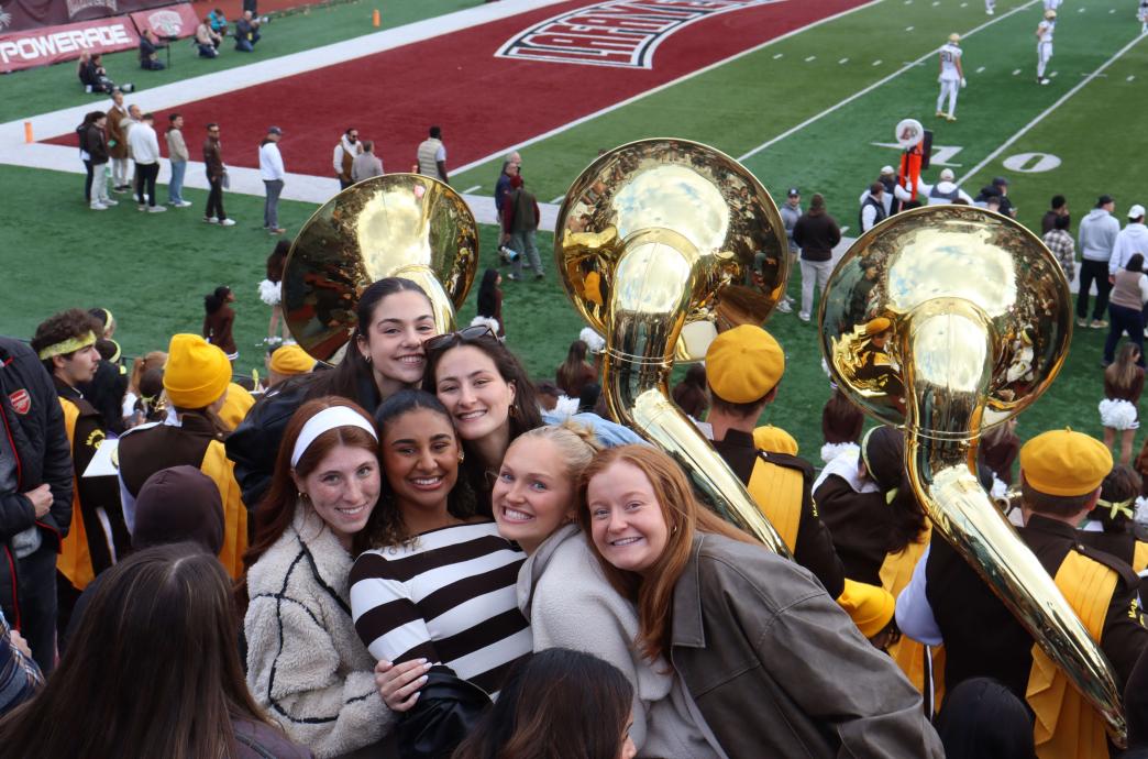 Six young women huddle together and smile. Three tubas poke up from the crowd behind them, and a bit of the field is visible.