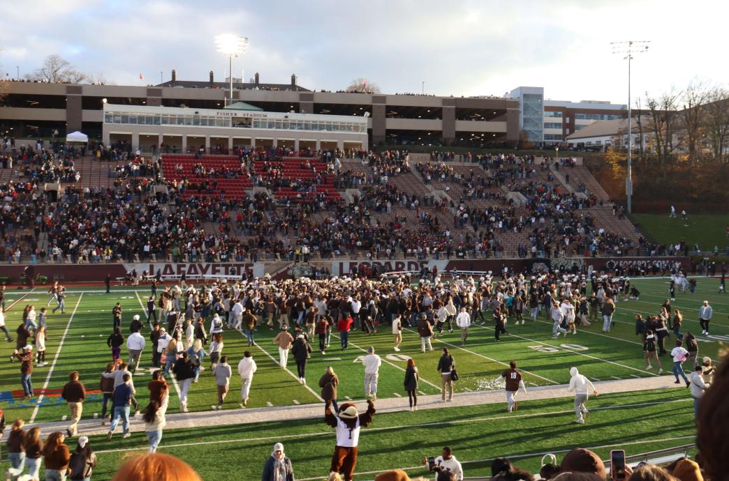 Dozens of fans swarm the field post Lehigh's win, cheering, while other fans fill the stands. Nearby, Clutch cheers.