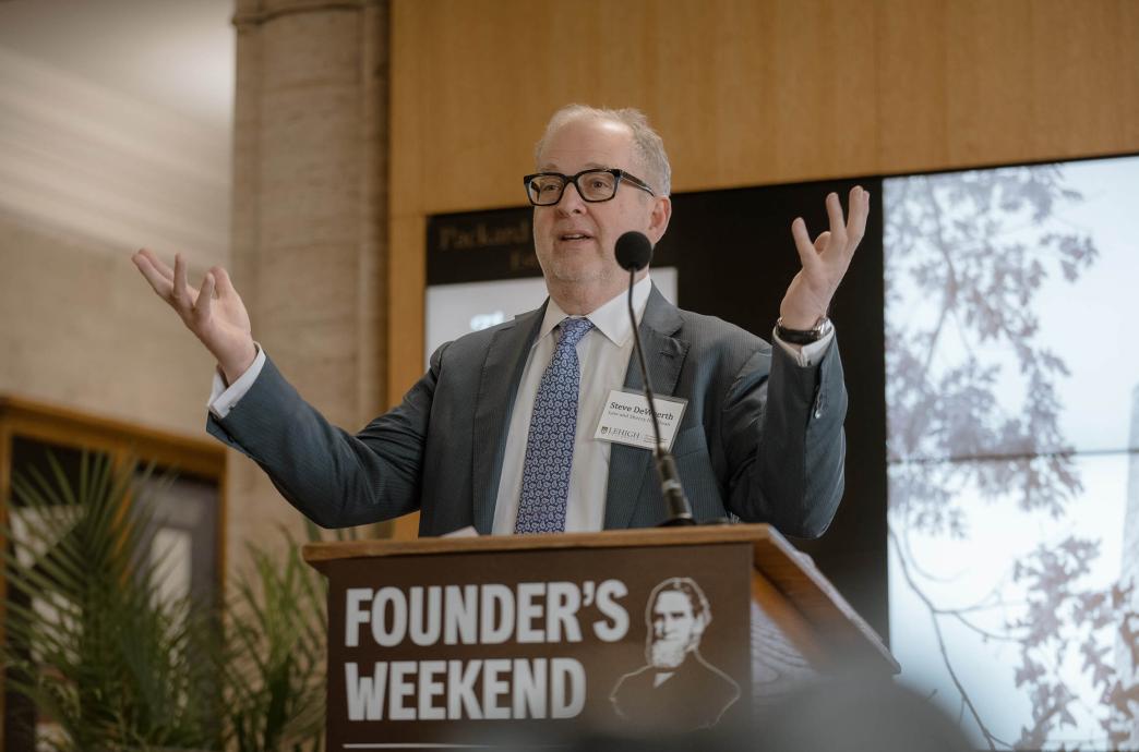 Dean DeWeerth stands at a podium wearing a gray suit and tie, speaking with his arms motioning up.