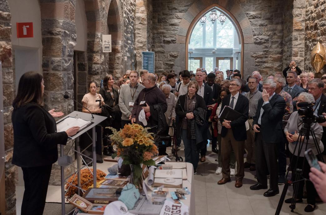 A crowd of people gather in the entry of the Clayton UC, listening to a woman speak at a podium with a table of time capsule items in front of her.