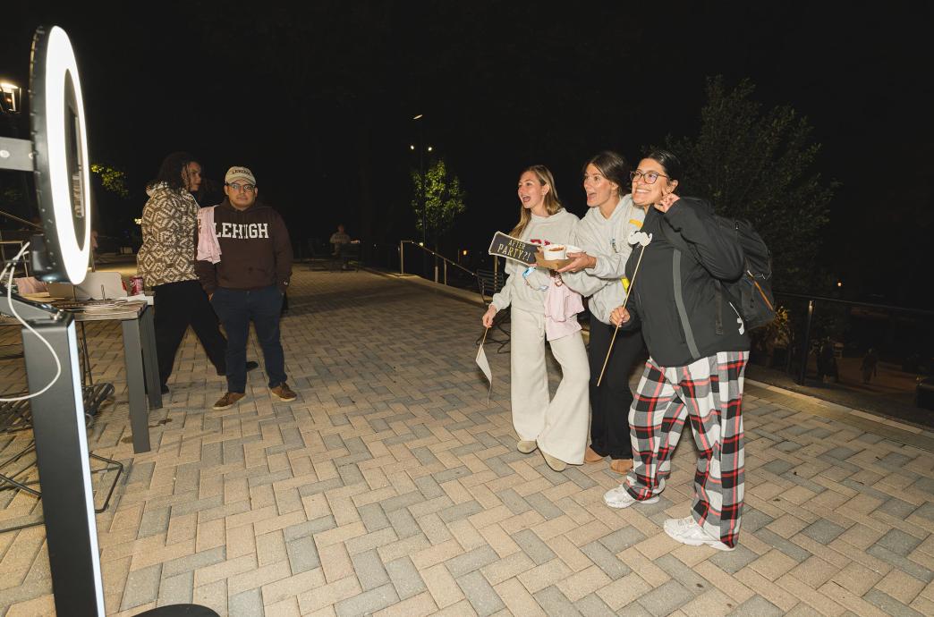 Three women wearing sweats and backpacks hold up photo booth props and pose in front of an open air photo booth ring light at night.