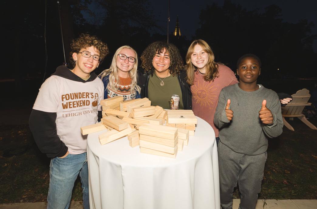 On a bar height round table with a white cloth, a stack of large jenga blocks are piled with five students posing for a photo behind it.