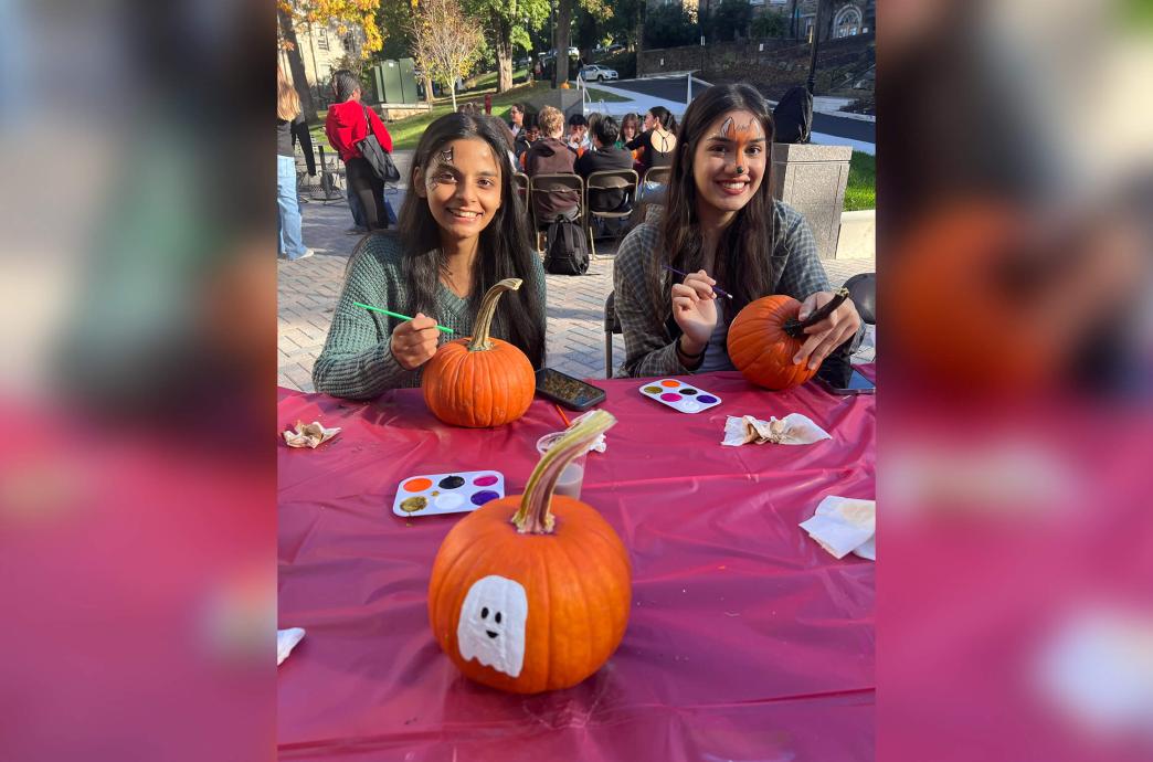 Two female students with their faces painted sit at a table with a pink table cloth, each painting pumpkins.