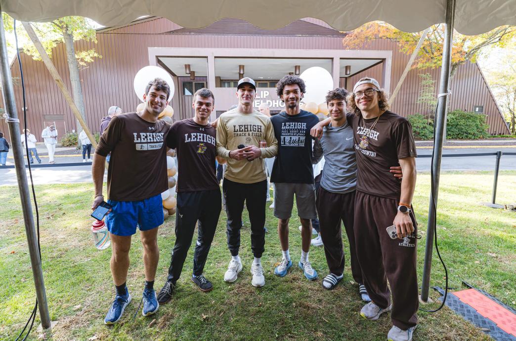 Standing at the edge of a white tent, six men pose for a photo wearing different style Lehigh track and field tshirts and athletic pants.