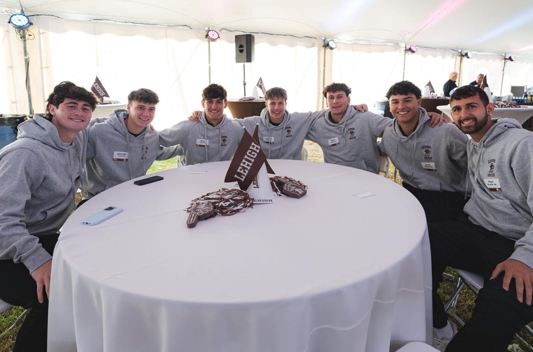 Under a large white tent, members of the Lehigh men’s soccer team sits at a white clothed round table, wearing matching gray pullover hoodies.
