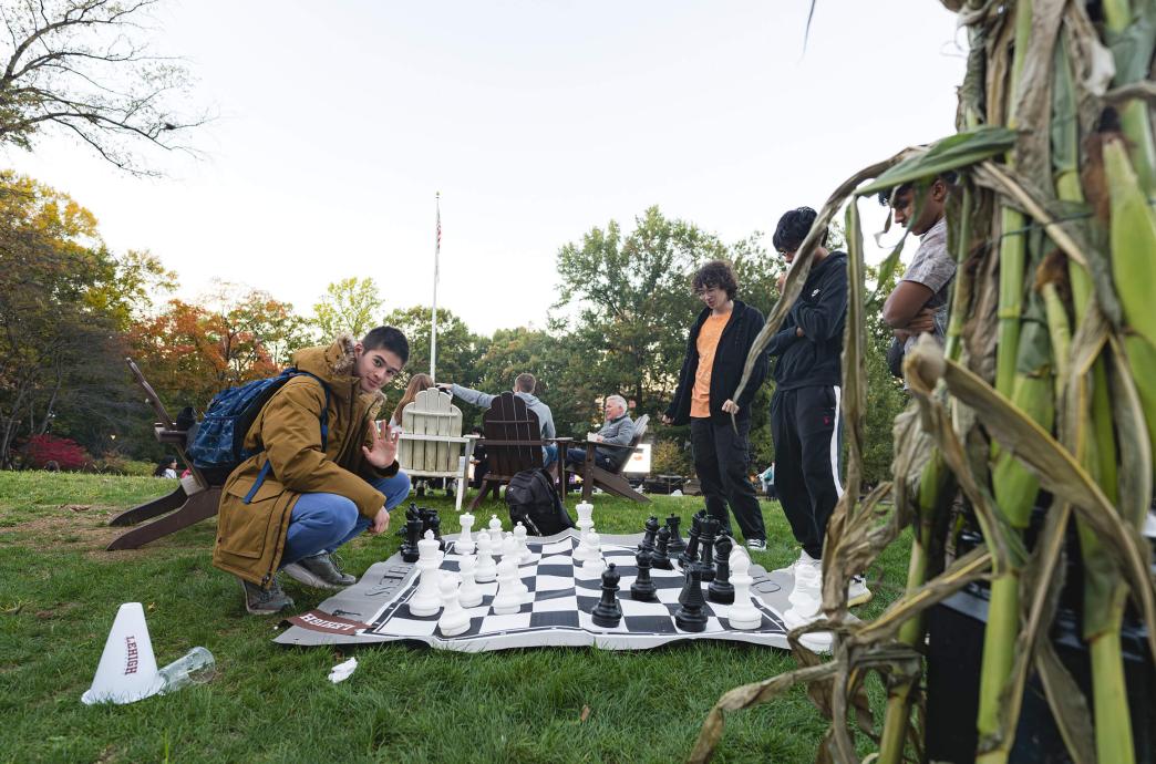 A student wearing a long brown jacket and a backpack squats next to a large, blanket-style chess set on the lawn of the UC with the flagpole in the distance.