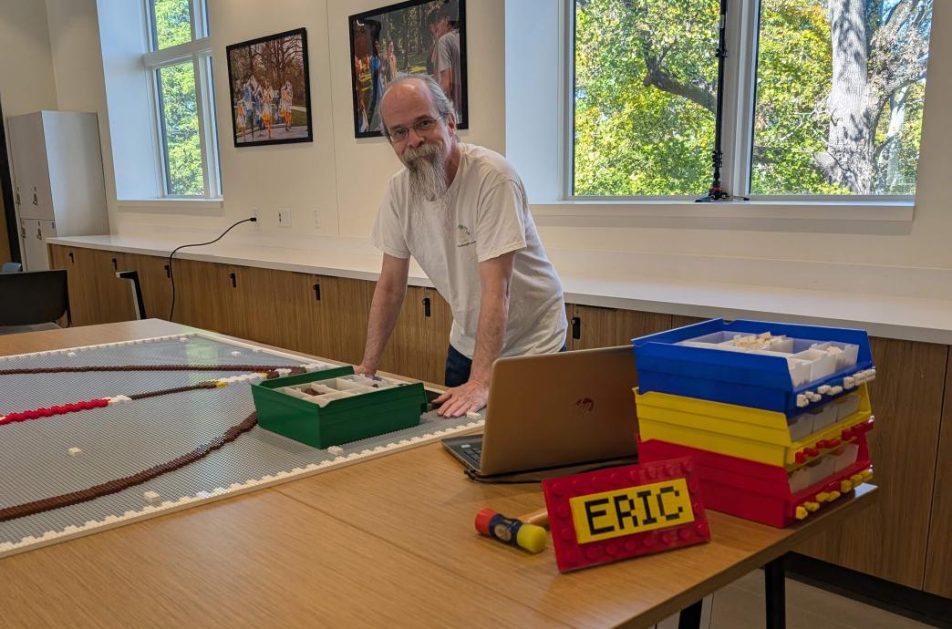 A man with a long white beard leans over a table covered with colorful boxes of legos and a brown shield project in progress.