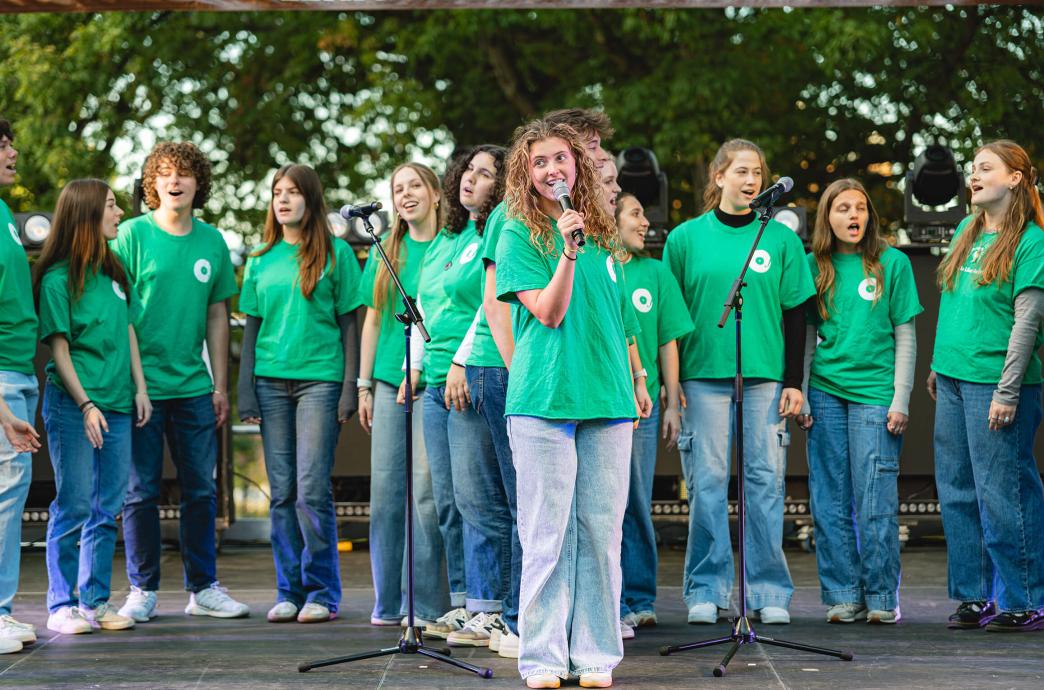 All dressed in denim pants and matching kelly green shirts, an acapella group sings on stage under a brown Lehigh banner, as one girl stands center holding a microphone.