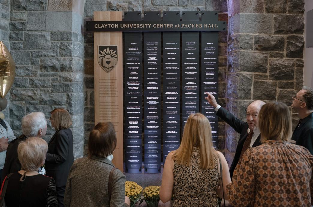 A man extends one hand to point to a recognition wall of names, hung on a stone wall with a crowd of people gathered around it.