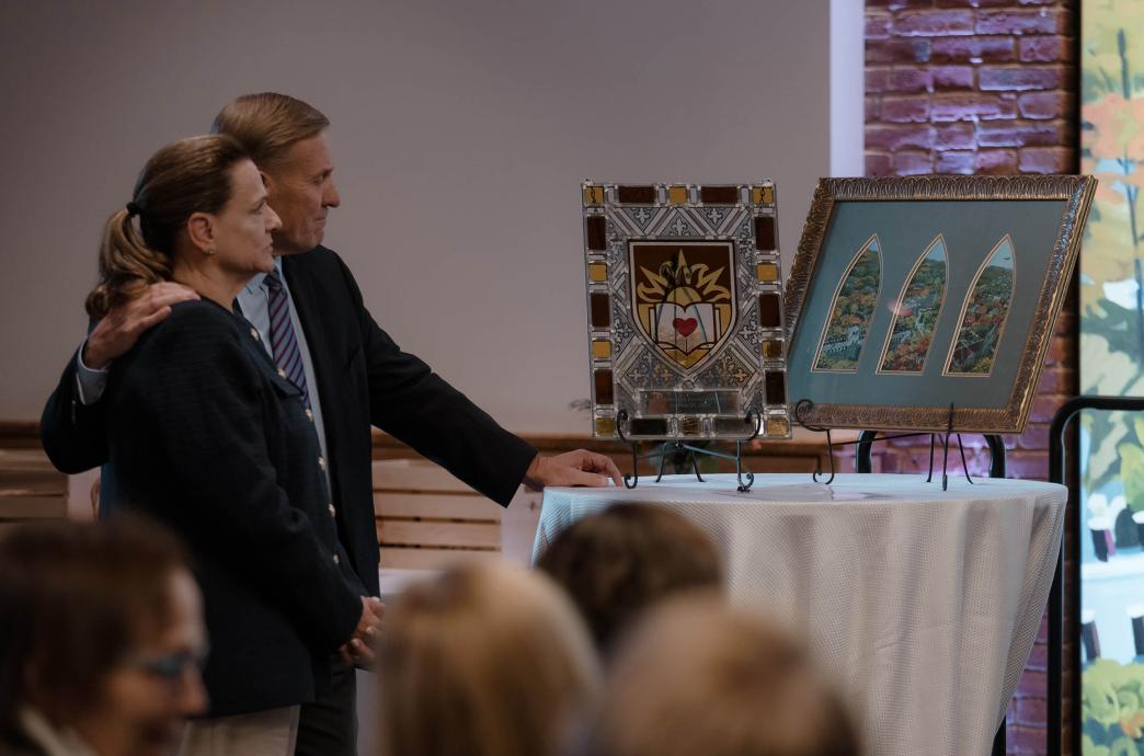 Kevin L. Clayton stands with his arm around his wife’s shoulders as they look at one framed piece of art and another stained glass window art piece resting on a table.