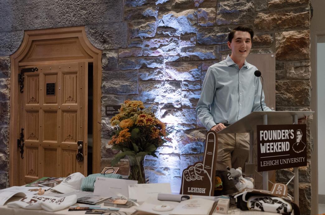 A student wearing a light blue button down shirt stands at a podium with a table of time capsule items to his right and a large wooden door propped open behind him.
