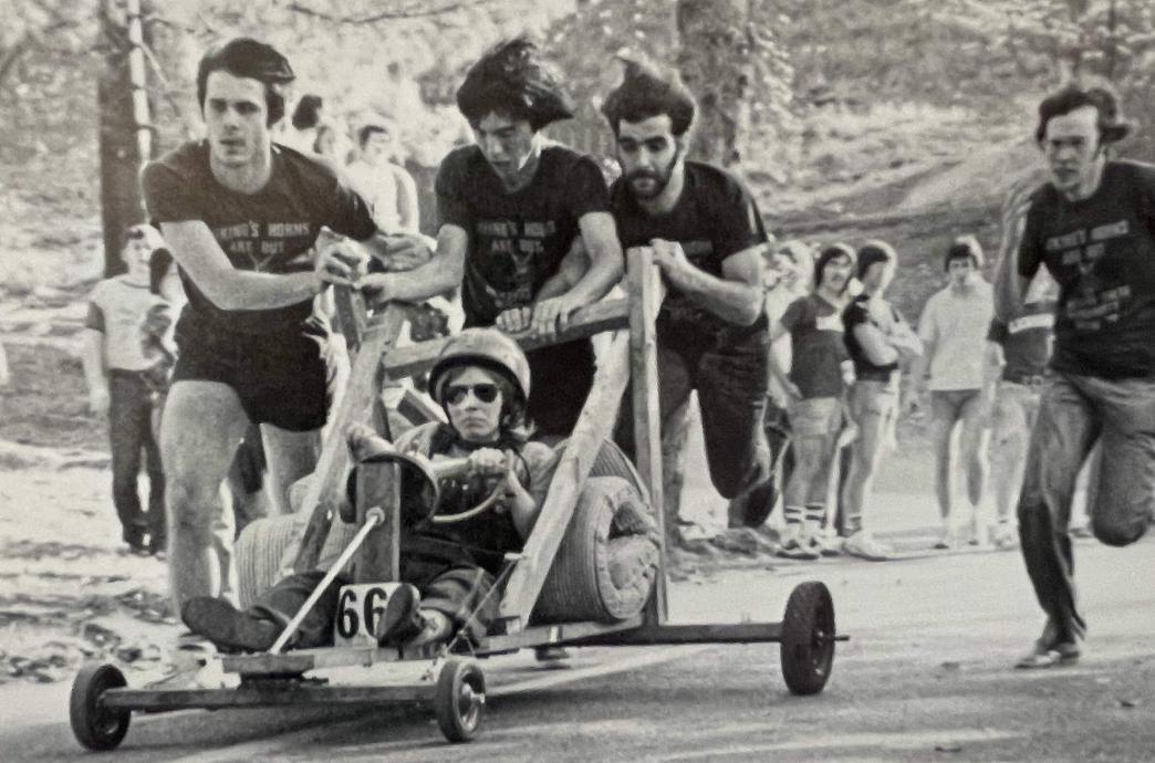 A black and white vintage photo of a hand made go-cart-style car, pusshed by men with a driver steering wearing sunglasses and a helmet.