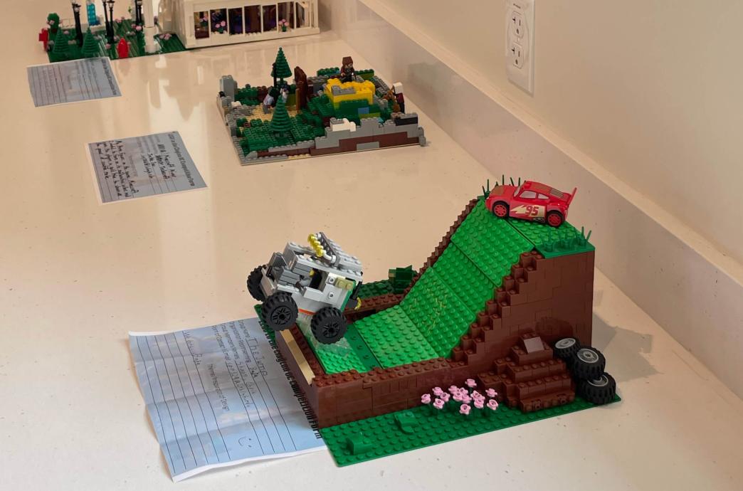Lego creations are lined up on a long, white table against a wall, with one in the foreground that shows a green hill with a truck jumping off the end.