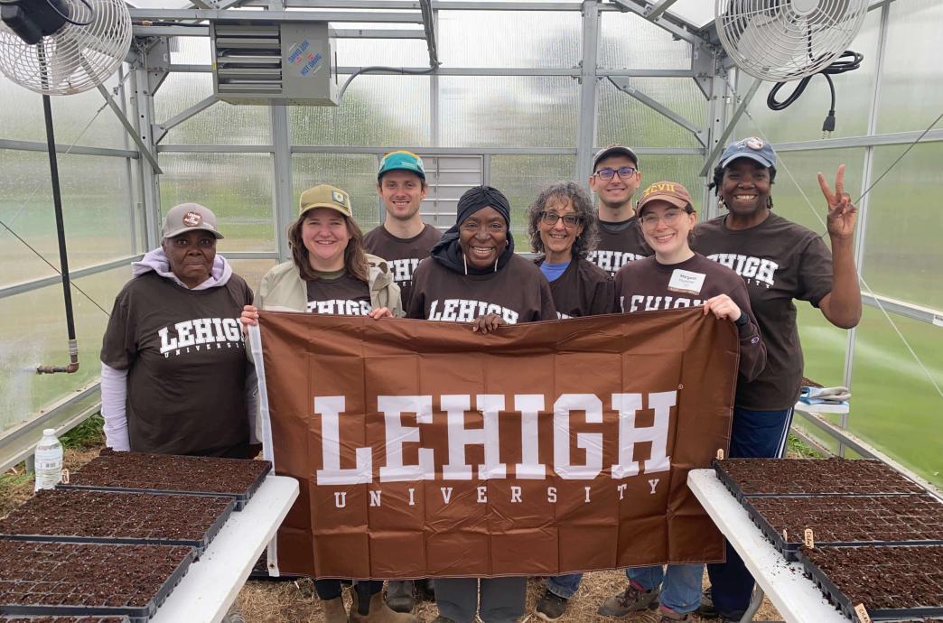 Eight volunteers stand in a greenhouse. They wear matching brown Lehigh shirts and hold a Lehigh banner up.