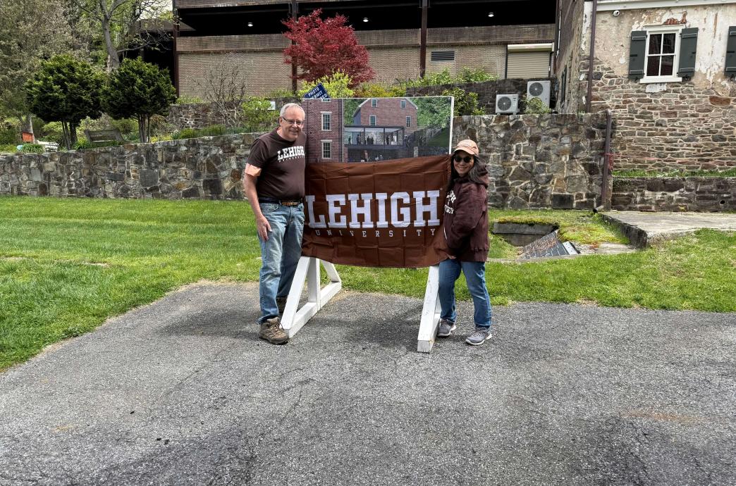 Two volunteers stand outside in historic Bethlehem. They wear matching brown Lehigh shirts and hold a Lehigh banner up.