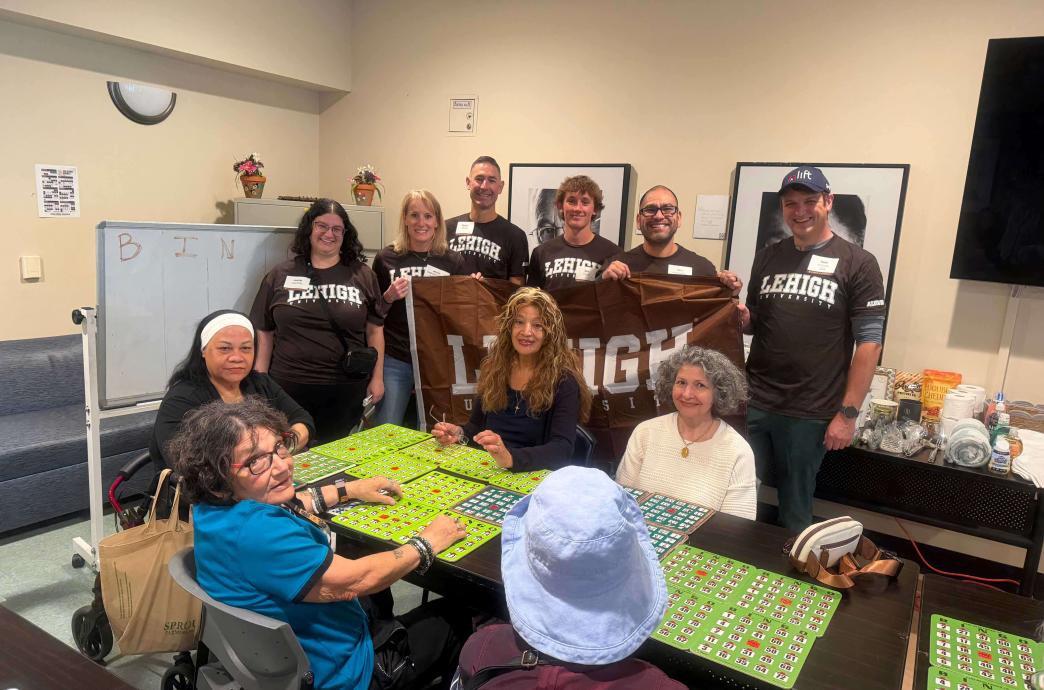 Six volunteers stand near seniors playing bingo. The volunteers wear matching brown Lehigh shirts and hold a Lehigh banner up.