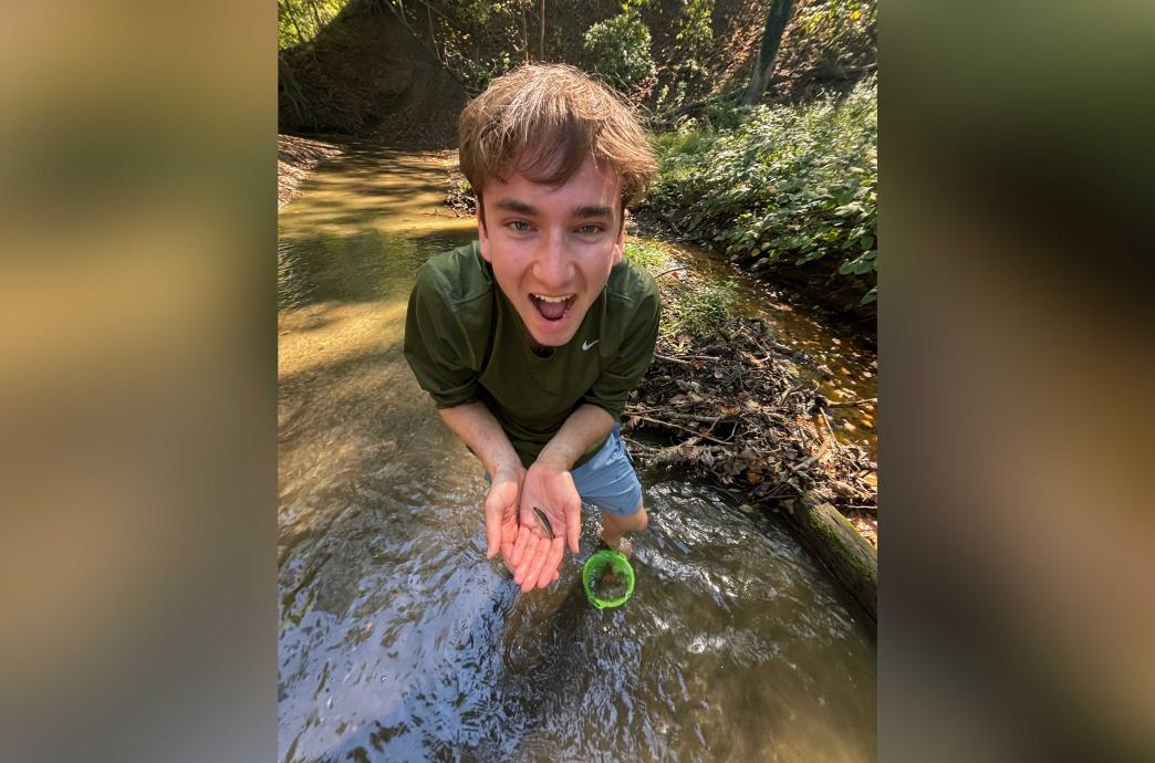 Garrett smiles widely while holding a tiny fish caught in the lake he stands in. A small green bucket is at his feet.
