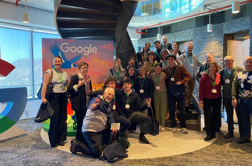 A group of students stand in Google Chile and smile. Behind them is a google sign with a blue bird on it.