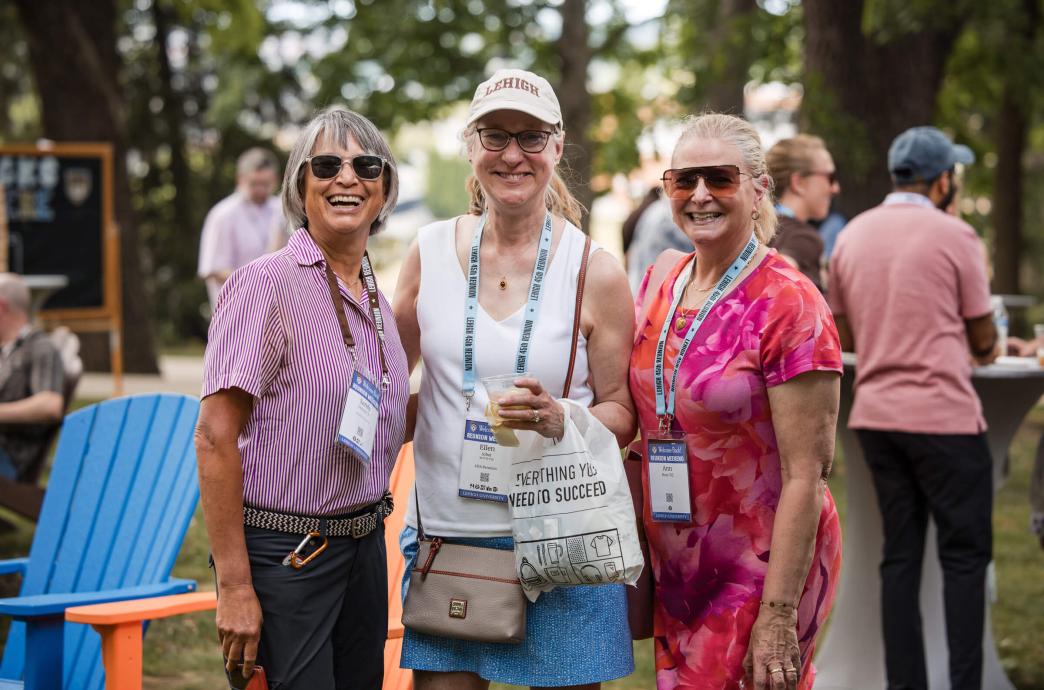 Three women stand together and smile at a Reunion event outside. They each wear blue lanyards with their names.