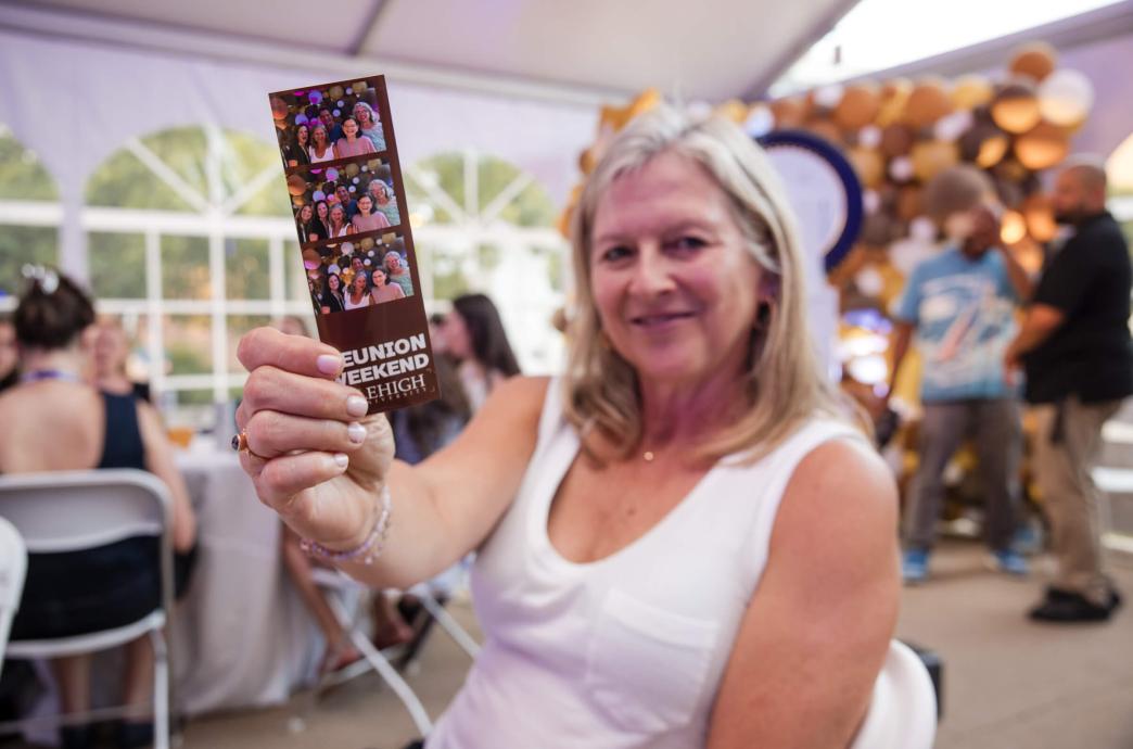 A woman sits near a photo booth and holds up her printed images. The photo strip is Lehigh branded and shows her with friends.