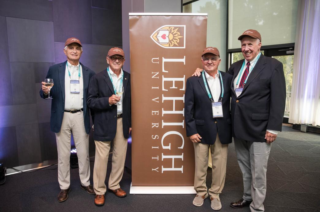 Four seasoned alumni stand near a Lehigh University banner. They wear matching Lehigh hats, suit jackets, and slacks. 