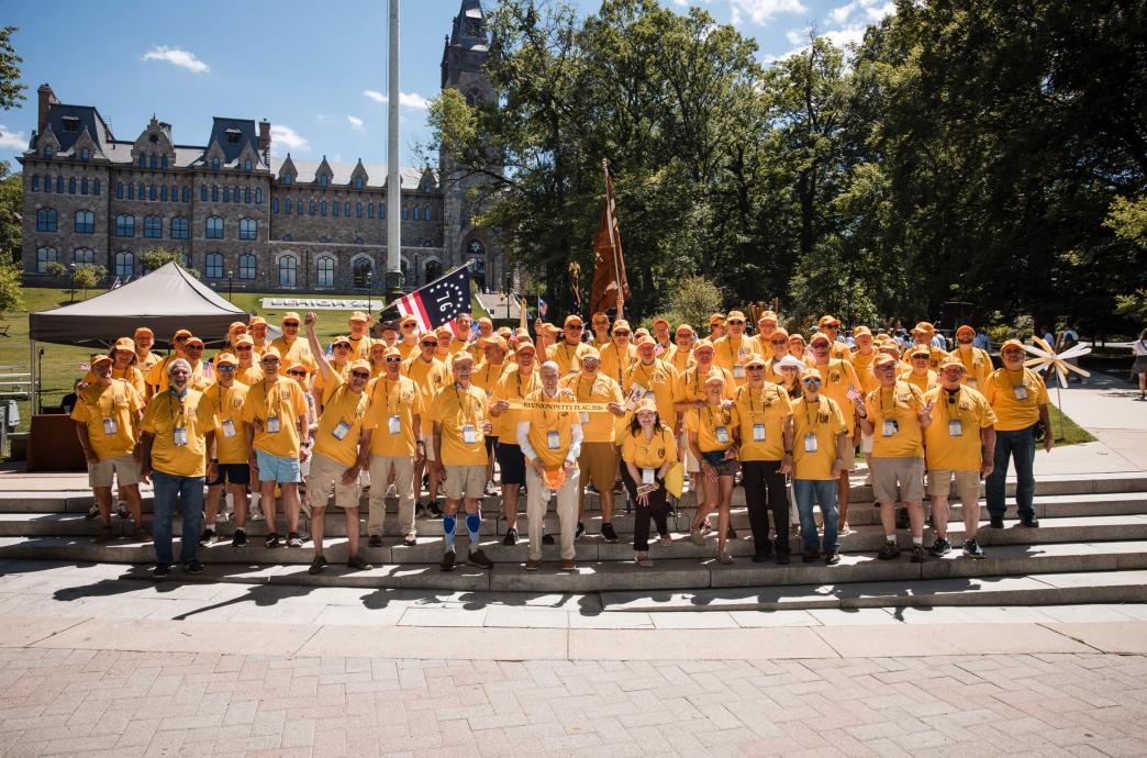 A large group of alumni wear bright yellow shirts and stand by the flag pole. They hold a small 'Reunion Petty Flag 2026' banner.