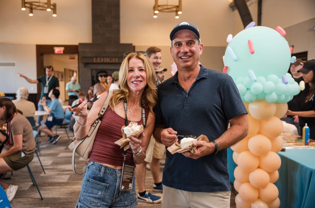 Two alumni stand together and smile while holding ice cream. An ice cream balloon is to the right of them near a toppings bar.