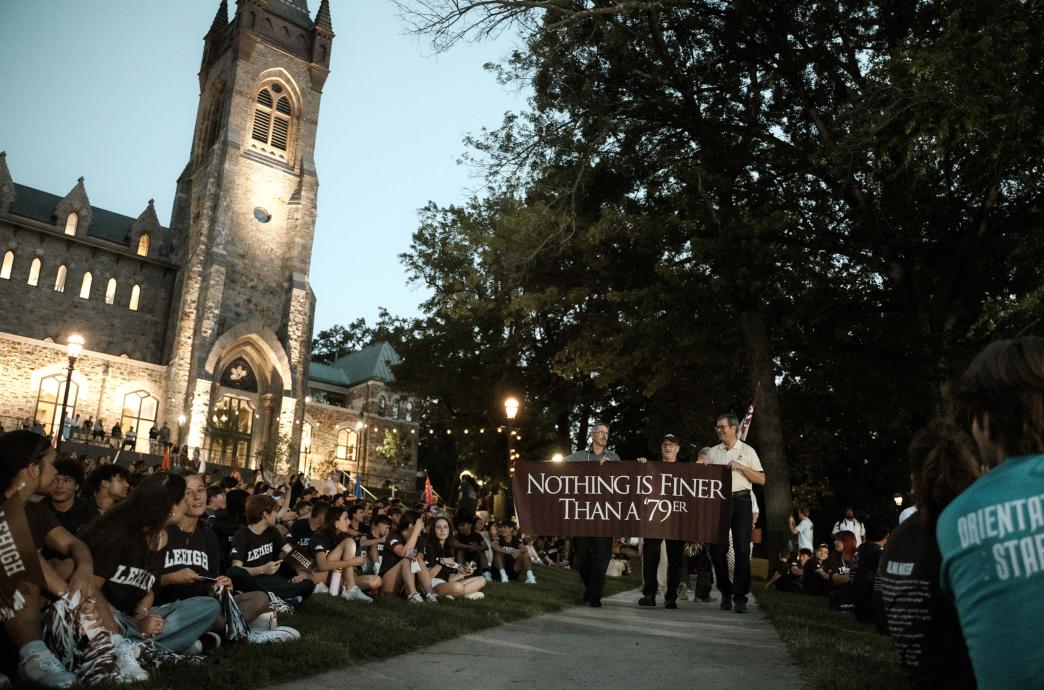 Three men hold a banner that reads “Nothing is finer than an ’79er” as they walk down a path lined with students in front of the University Center.