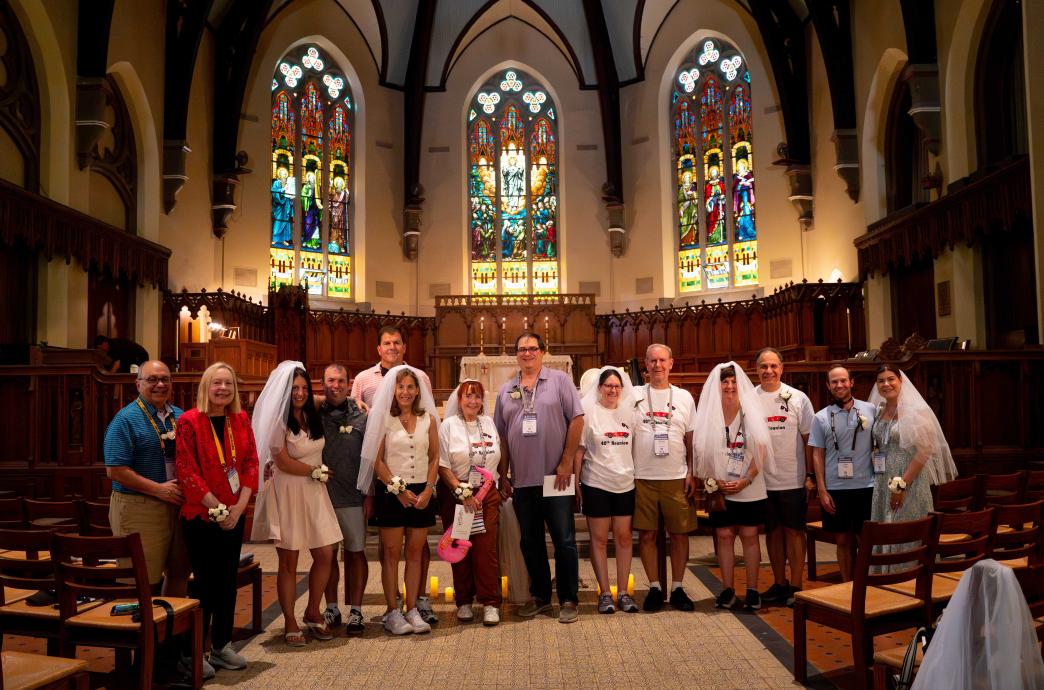 A group of alumni stand in Lehigh's chapel and smile. Many of the women wear veils and a corsage on their wrists. 