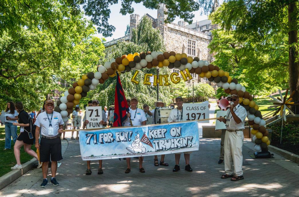 A group of alumni hold a Class of 1971 sign and smile. They stand below a balloon arch on campus and wave their class flag.