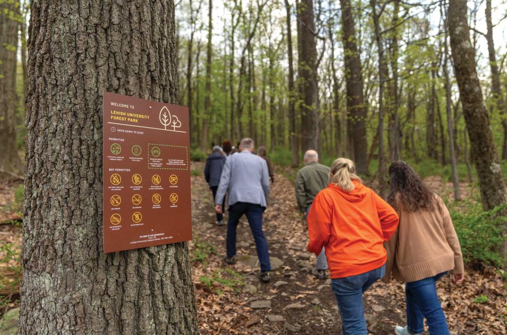 A group of people ascend a hillside trail where a tree with a sign shares information on a new park.