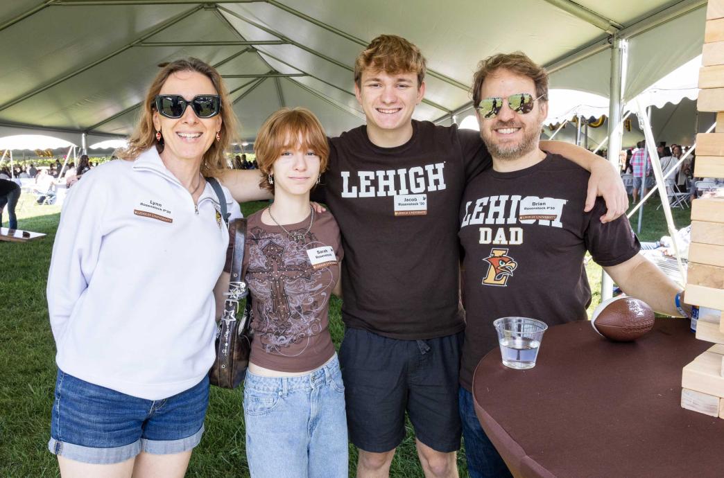 A family stands together under a tent while tailgating. They stand beside a Jenga tower. 