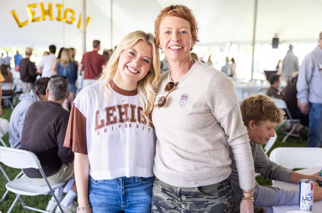 Two women wear Lehigh merch and smile under a tent while tailgating. Lehigh balloons are visible behind them.