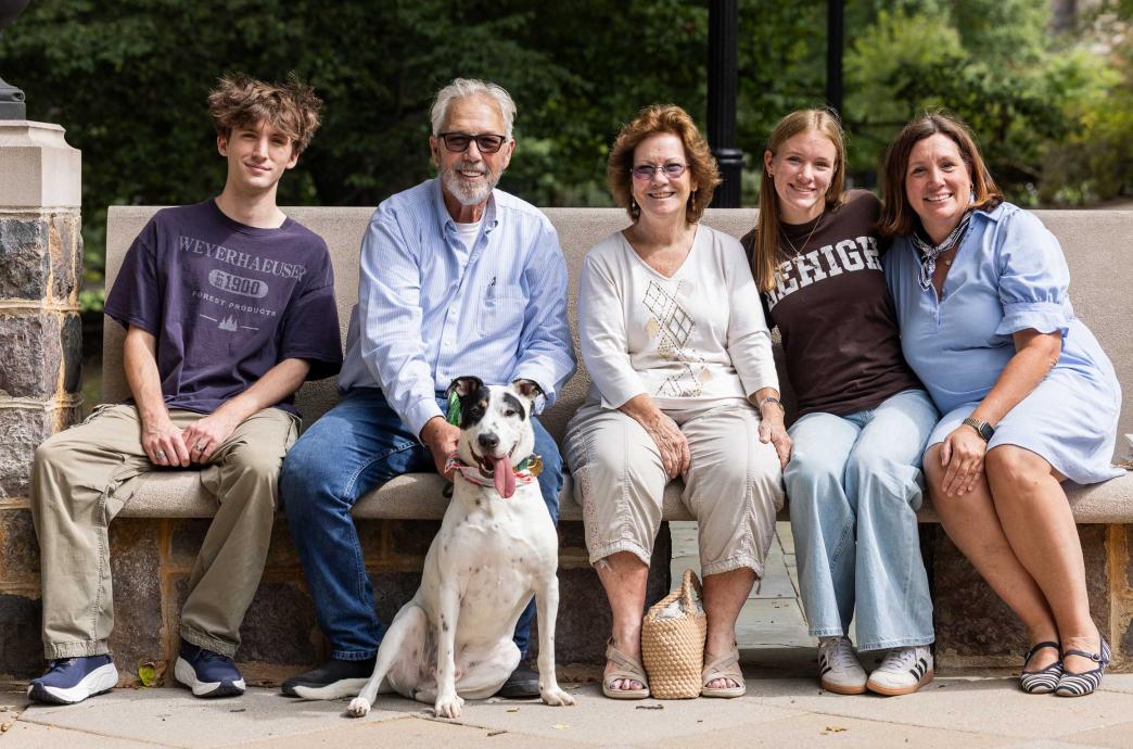 A family sits together on a bench and smiles. A black and white dog sits in front of them.