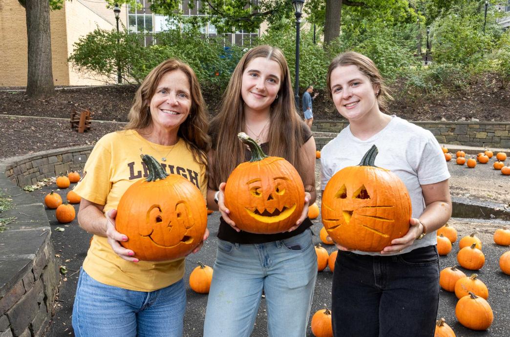 Three women stand together and hold up pumpkins with silly faces carved into them.