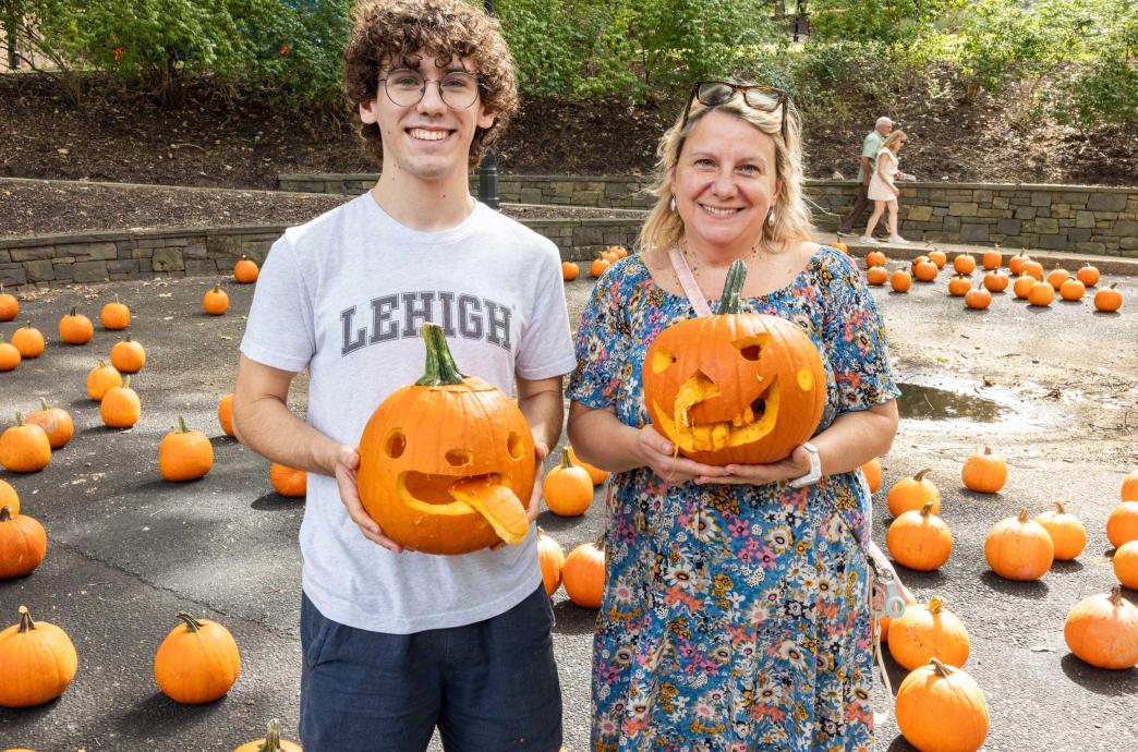 A young man in Lehigh merch stands alongside a woman in a floral dress. They both hold up carved pumpkins.