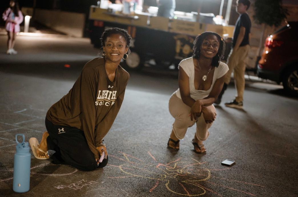 Two young women kneel on the blacktop while holding chalk and drawing figures.