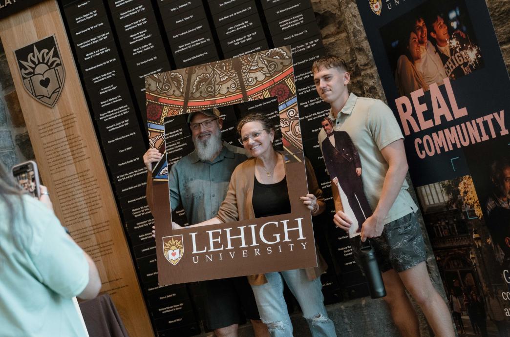 A family smiles while someone snaps a photo. The parents hold up a Lehigh frame while the student holds a photo of Asa Packer.