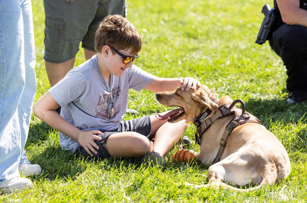 A young boy sits cross-legged on the grass and leans forward to pet Grace, an LUPD golden retriever. 