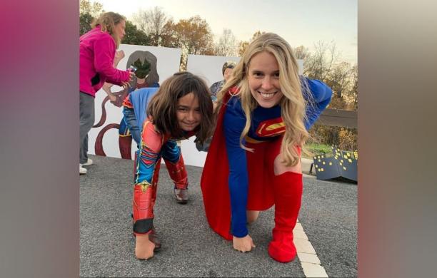 Faith Roncoroni dressed as Supergirl kneeling in a hero pose with a child in a Captain Marvel costume
