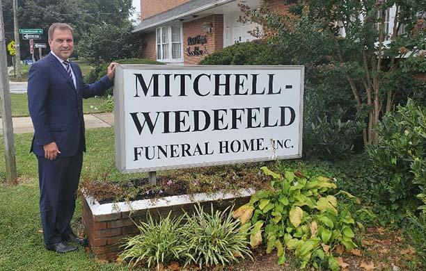 Jack Mitchell in a blue suit stands next to the funeral home sign that bears his name