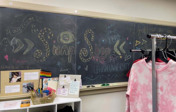 A blackboard covered with colorful doodles and says "Welcome to the Swap Shop, Lehigh University" beside a rack of clothes